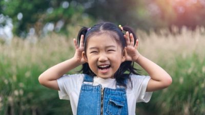 a small girl smiles and holds her ears as if listening to a noise in an outdoor setting.