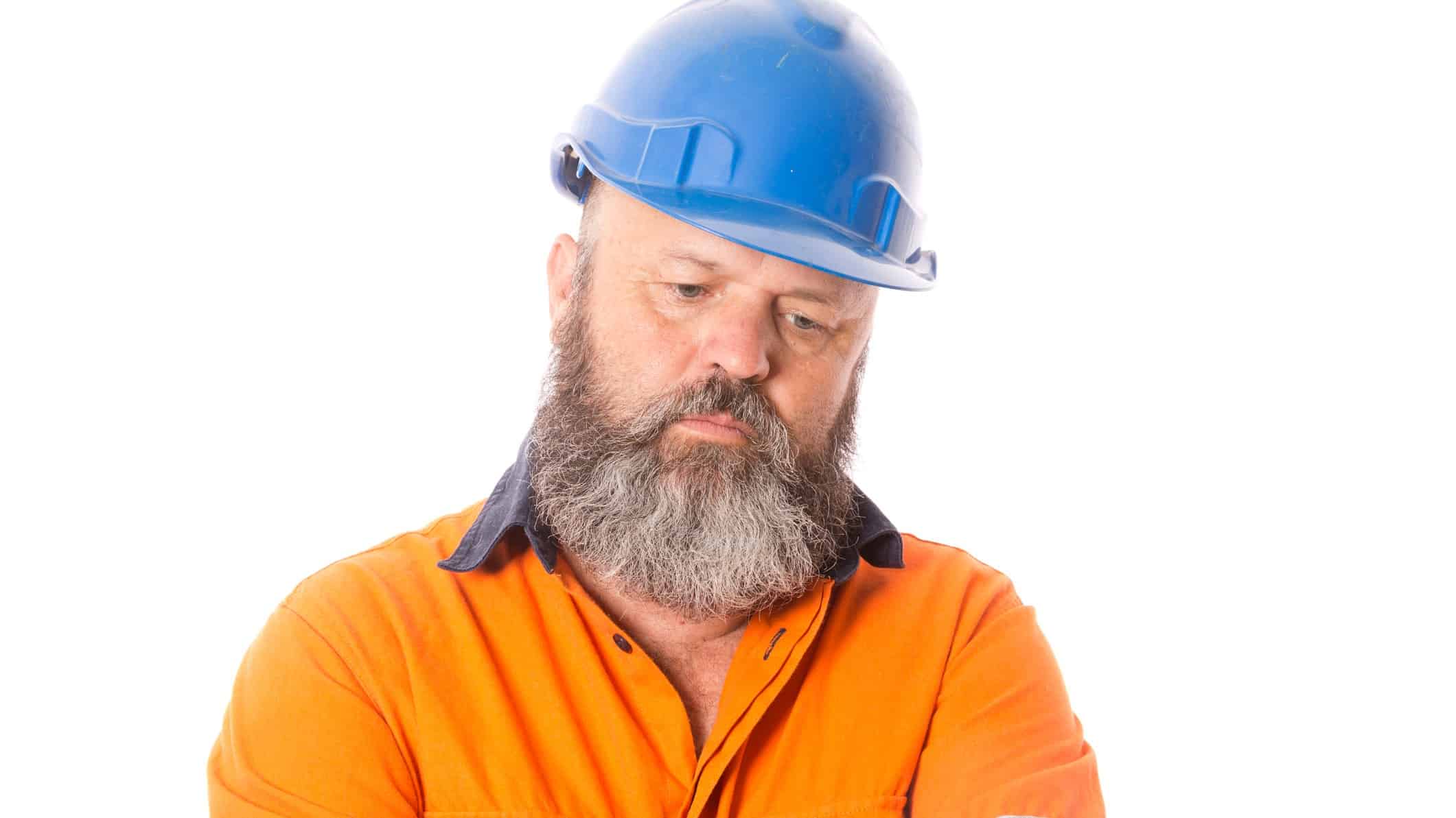 a man in high visibility shirt and hard hat with full beard looks downcast with eyes lowered as though he is disappointed or sad.