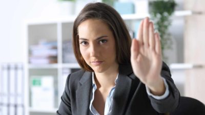 a woman wearing a dark business suit holds her hand up in a stop gesture while sitting at a desk. She has a sombre look on her face.