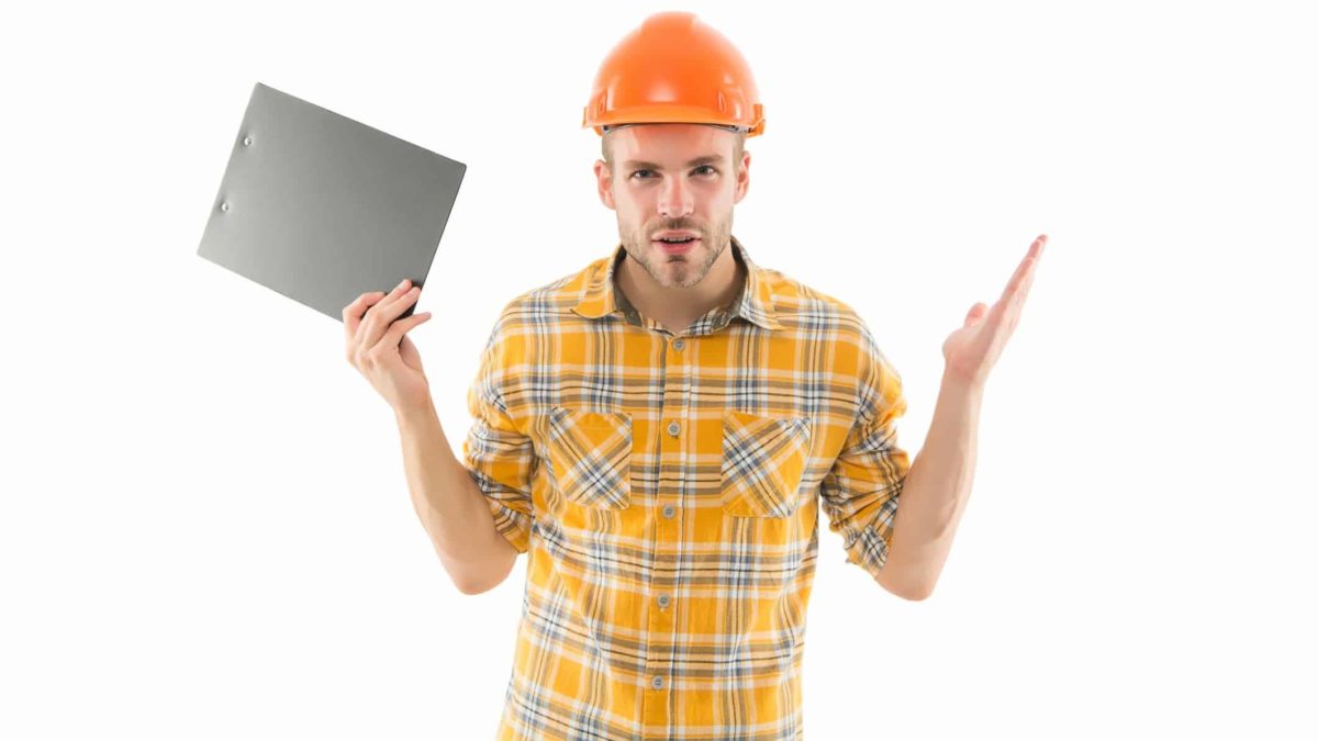 a man in a hard hat and checkered shirt holds paperwork in one hand as he holds his hands upwards in an enquiring manner as though asking a question or exasperated by uncertainty.