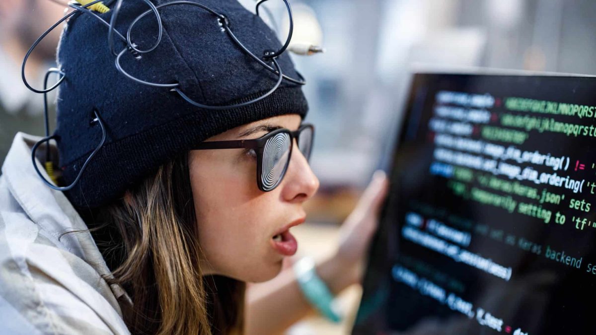 a woman wearing a close-sitting hat featuring wires and thick computer screen glasses clutches her computer monitor and looks shocked and disturbed as she reads old-fashioned computer text from the screen.
