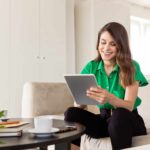 A happy woman smiles as she looks at a tablet in a room with green plant life around her.