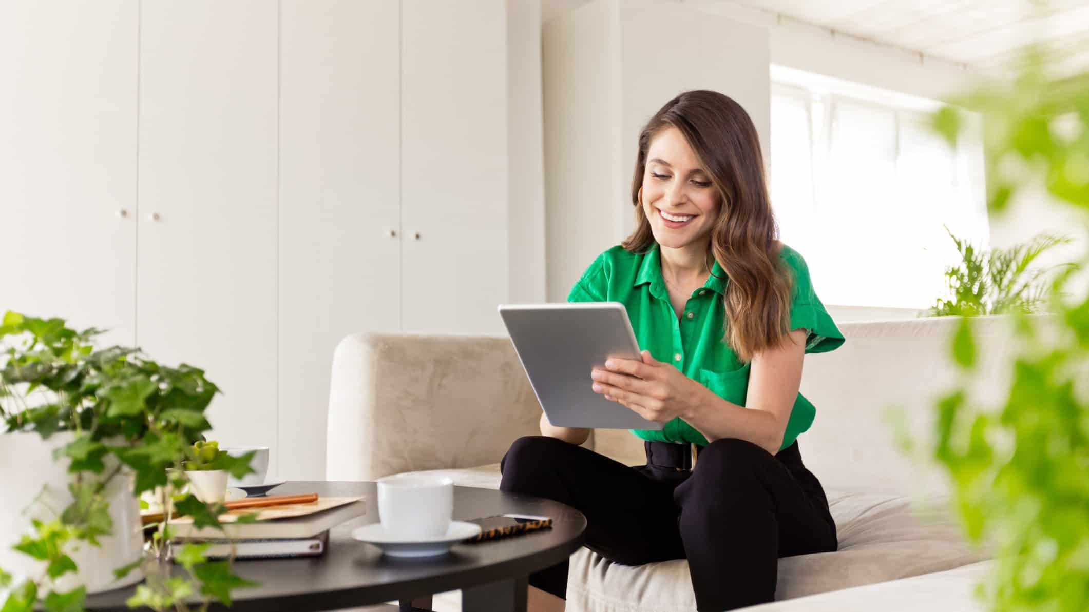 a happy woman smiles as she looks at a tablet in a room with green plantlife. She is also wearing a green shirt.