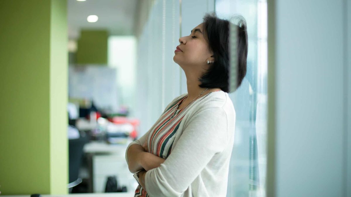 a woman leans her back on the glass of an office tower with her arms folded and her eyes closed as if digesting bad news.