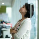 a woman leans her back on the glass of an office tower with her arms folded and her eyes closed as if digesting bad news.
