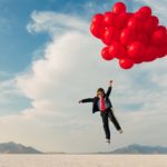 A boy dressed in a business suit and old-fashioned flying helmet and goggles is lifted by a bunch of red helium balloons over a barren desert landscape.