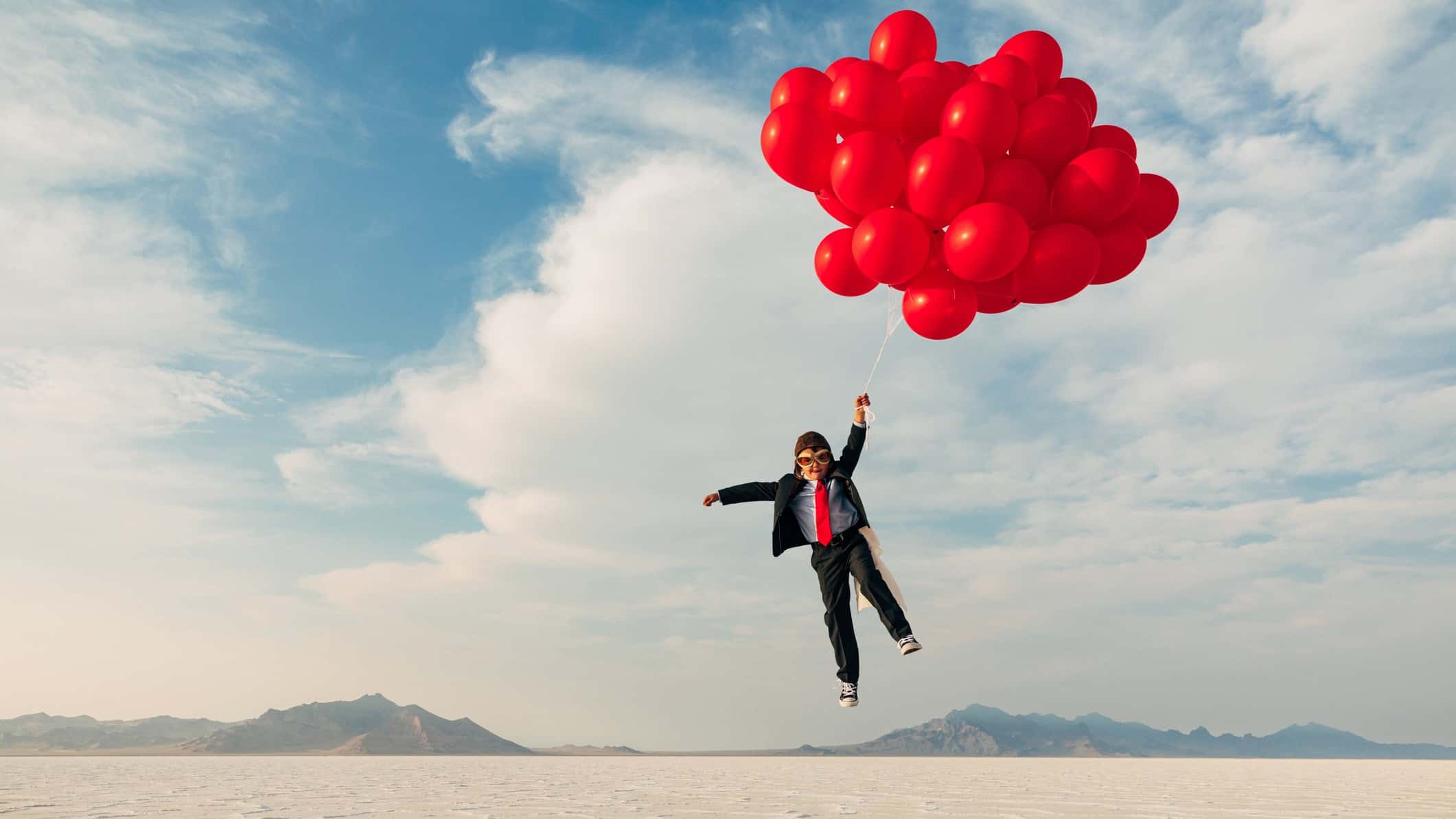 a boy dressed in a business suit and old fashioned flying helmet and goggles is lifted by a bunch of red helium balloons over a barren desert landscape.