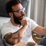 A hipster-looking man with bushy beard and multiple arm tattoos sits on the floor against a sofa reading a tablet with his hand on his chin as though he is deep in thought.