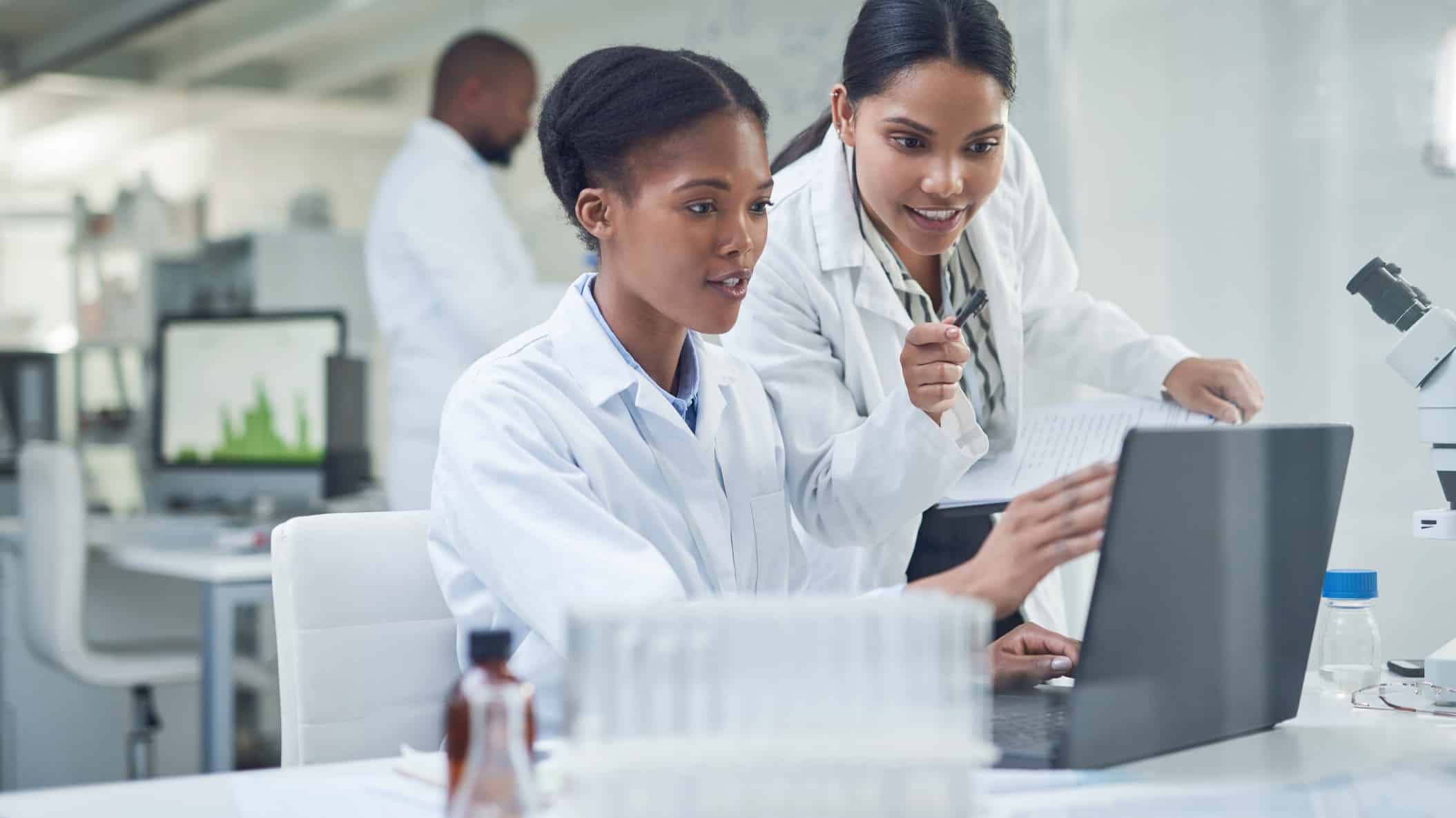 Two Archer Materials lab assistants wearing white coats discuss results they see on a computer screen
