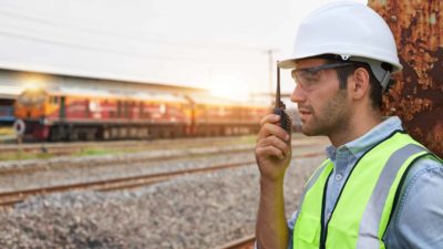 a man in hard hat and high visibility vest talks into a walky-talky device in the foreground of a freight train at a railway yard.