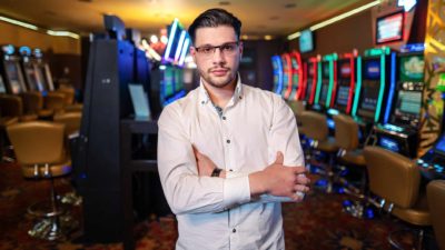a man stands with his arms folded in front of banks of unused poker machines in a darkened gaming room.