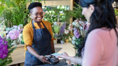 A smiling market stall holder selling flowers holds out a payment machine to a customer who hovers her telephone over it to pay via Zip