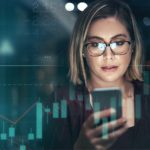 a woman checks her mobile phone against the background of illuminated share market boards with graphs and tables.