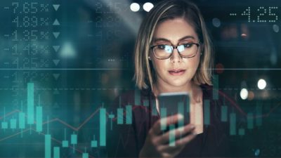 a woman checks her mobile phone against the background of illuminated share market boards with graphs and tables.