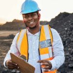 a man with a hard hat and high visibility vest stands with a clipboard and pen in front of a large pile of rock at a mining site.