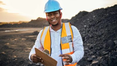 a man with a hard hat and high visibility vest stands with a clipboard and pen in front of a large pile of rock at a mining site.