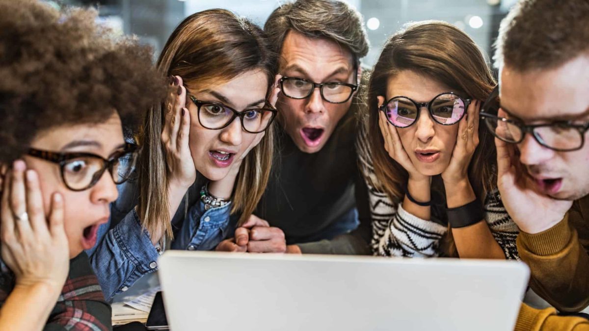 A group of people gathered around a laptop computer with various expressions of interest, concern and surprise on their faces. All are wearing glasses.