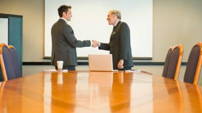 two men in suits shake hands at the top of a shined wood boardroom table.