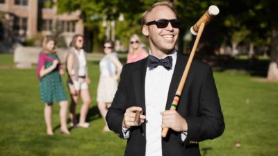 a man wearing a dinner suit holds a cigar and a croquet mallet on a rolling green lawn with a group of women and a mansion in the background.