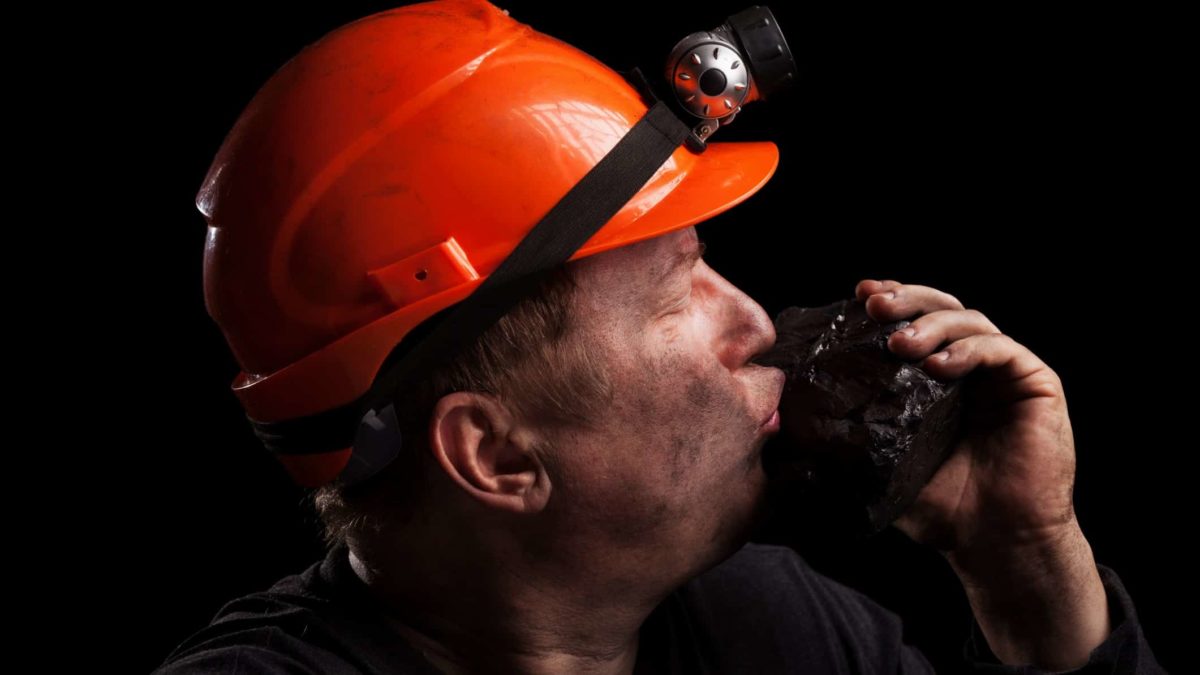 a coal miner in hard hat with a light on it kisses a large lump of coal that he is holding in his hand.