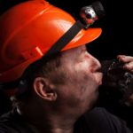 a coal miner in hard hat with a light on it kisses a large lump of coal that he is holding in his hand.