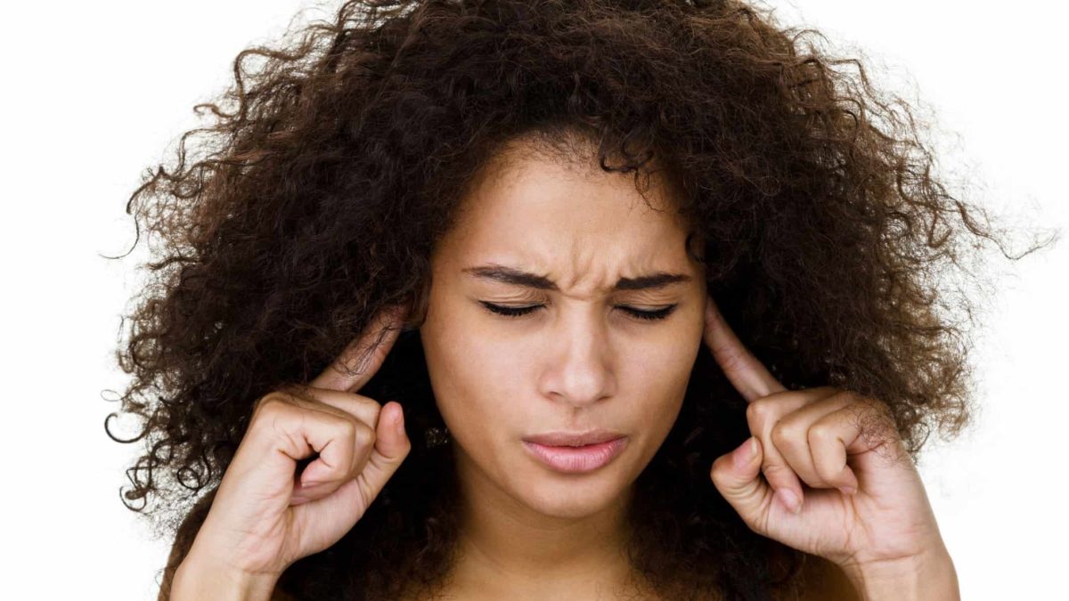 a woman puts her fingers in her ears with a pained expression on her face with her eyes closed as though trying to block hearing bad news or an unpleasant loud noise.