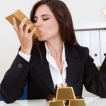 A woman in a business suit sits at her desk with gold bars in each hand while she kisses one bar with her eyes closed. Her desk has another three gold bars stacked in front of her. symbolising the rising Northern Star share price