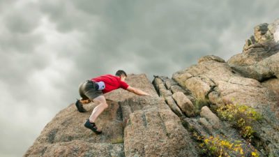 a climber scales a sheer rock cliff face reaching out for a handhold with foreboding grey clouds gathering in the sky above him.