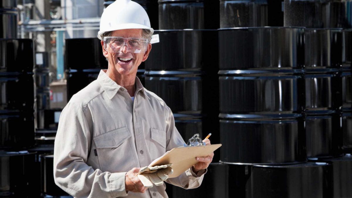 a man stands in overalls and a hardhat with a clipboard in front of stacked black oil drums at an oil industry site.