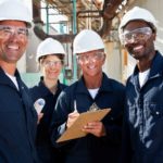 a group of four engineers stand together smiling widely wearing hard hats, overalls and protective eye glasses with the setting of a refinery plant in the background.