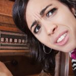 A woman with a pained and confused look on her face twiddles the dial on an old fashioned radio receiver while leaning in to listen to it.