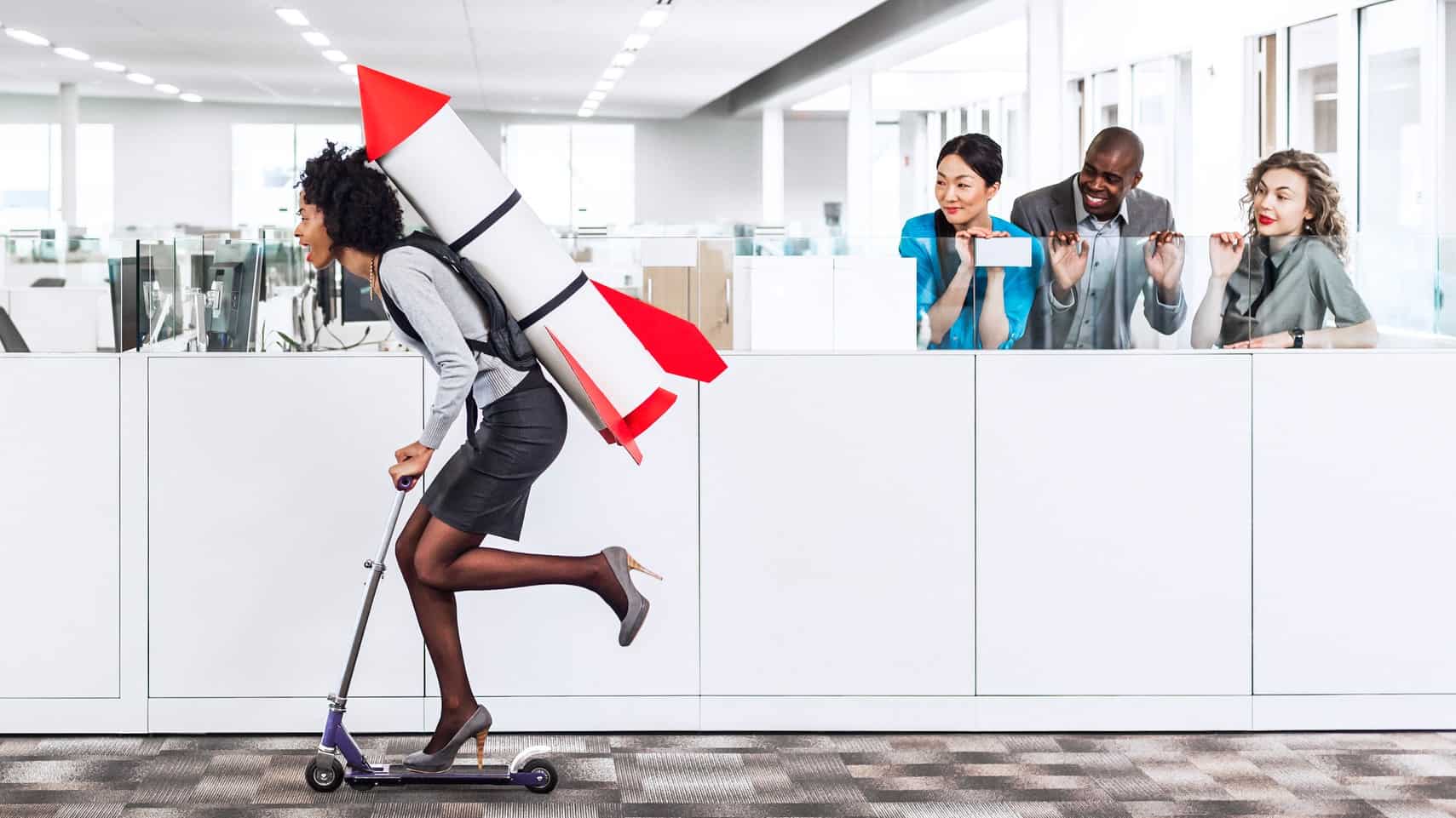 A woman rides through an office on a scooter with a rocket strapped to her back as colleagues cheer.