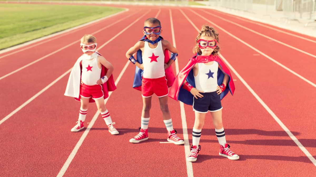 three children wearing superhero costumes, complete with masks, pose with hands on hips wearing capes and sneakers on a running track.