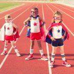three children wearing superhero costumes, complete with masks, pose with hands on hips wearing capes and sneakers on a running track.