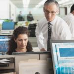 A group of market analysts sit and stand around their computers in an open-plan office environment.