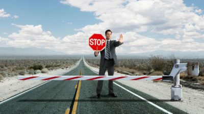 a man in a suit holds up a hand and a stop sign at a roadblock positioned over a bitumen road .