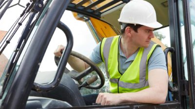 A man in high visibility vest and hard hat at the wheel of heavy mining machinery looks backwards out of the cabin window.