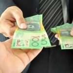 A man in a business suit whose face isn't shown hands over two australian hundred dollar notes from a pile of notes in his other hand to an outstretched hand of another person.