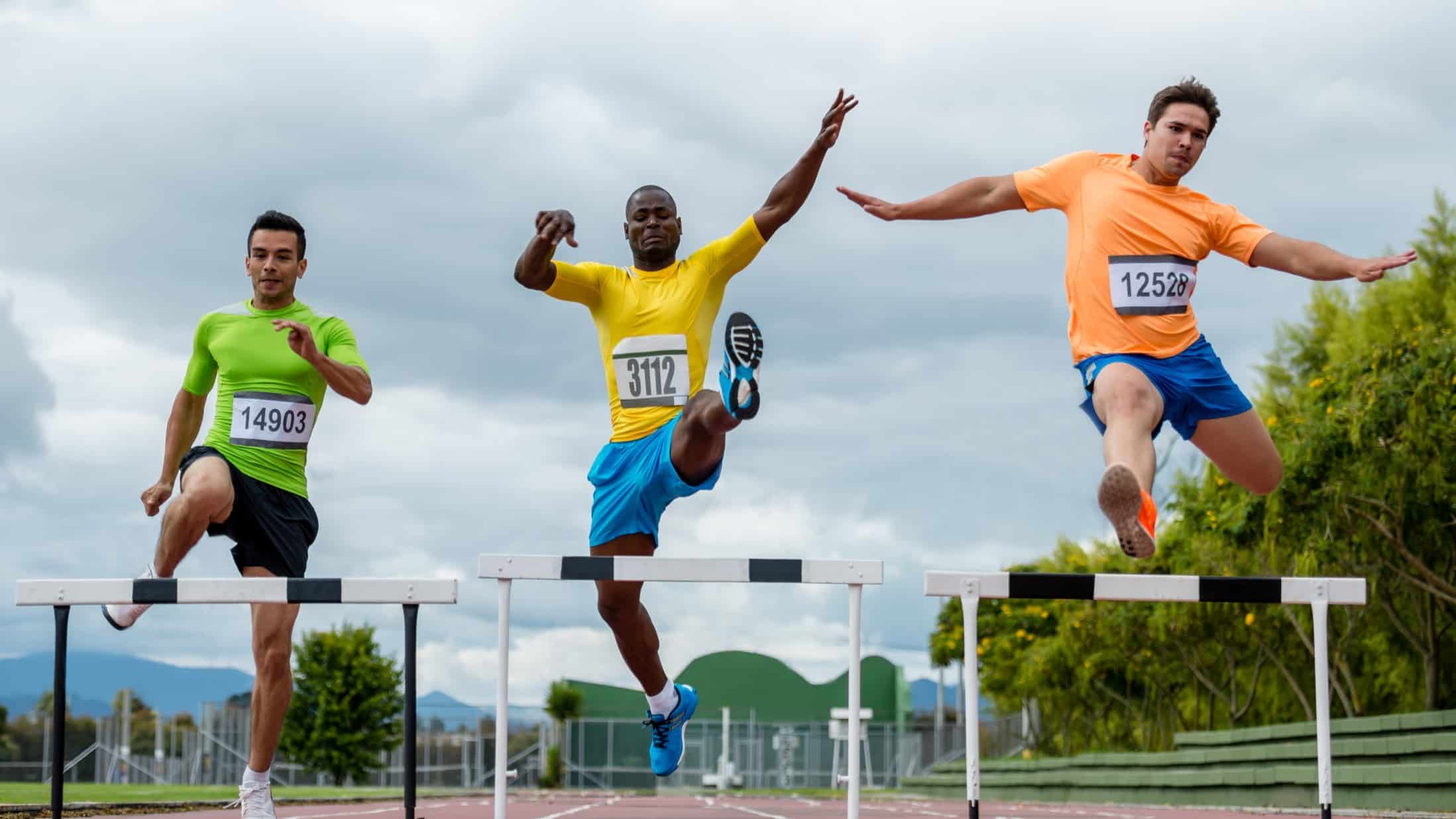 three people wearing athletic numbers and outfits jump over hurdles on a running track.