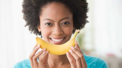 a woman holds a banana up to her face, mirroring her own smile as she holds the banana with two hands.