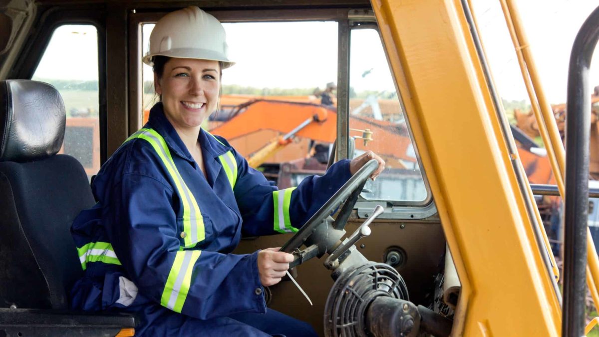 A GWR Group female employee in a hard hat and overalls with high visibility stripes sits at the wheel of a large mining vehicle with mining equipment in the background.