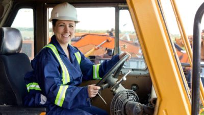 A female employee in a hard hat and overalls with high visibility stripes sits at the wheel of a large mining vehicle with mining equipment in the background.
