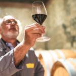 A wine technician in overalls holds a glass of red wine up to the light and studies it.