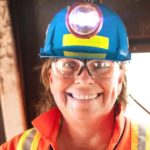 a woman wearing full miner's uniform, including a hard hat with lamp, high visibility overalls and vest, smiles in front of mining equipment.