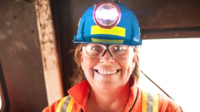 a woman wearing full miner's uniform, including a hard hat with lamp, high visibility overalls and vest, smiles in front of mining equipment.