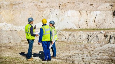 a group of three miners in hard hats and high visibility vests confer at a rocky mining site.