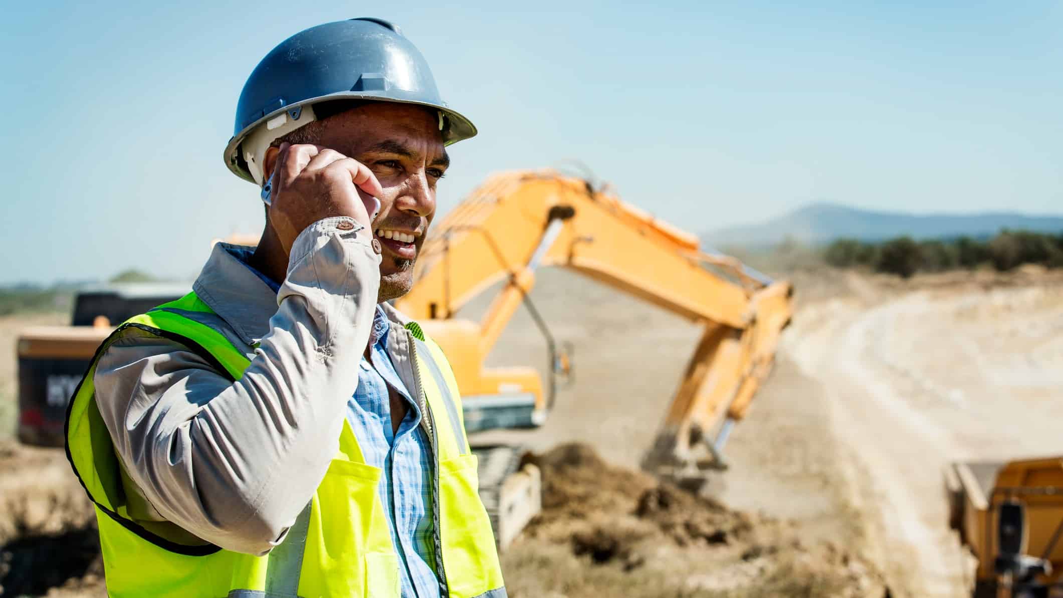 a man in hard hat and high visibility vest speaks on his mobile phone in front of a digging machine with a heavy dump truck vehicle also visible in the background.