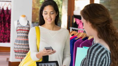 a woman looks at her phone while making a transaction at the counter of a store where racks of clothing can be seen in the background.