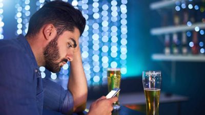 a man sits at a bar with a half full glass of beer and looks sadly into his mobile phone while propping his head on his hand with his elbow resting on the bar.