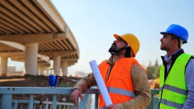 two road construction workers in hard hats and high visibility vests look up at an elevated section of road.